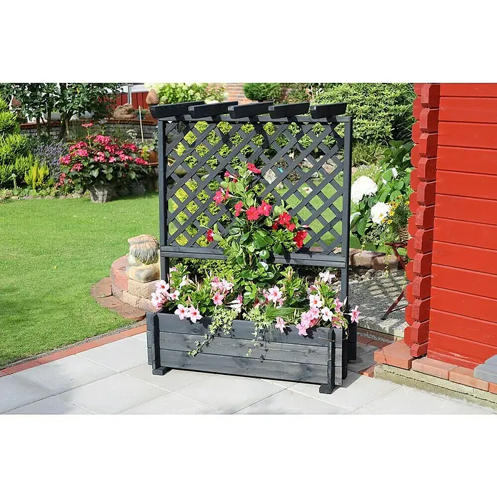 Promadino Pflanzkasten mit Pergola - Außenmaß (L x B x H): 140 x 64,5 x 102 cm, Holz, Anthrazit - Ein stilvoller, dunkelgrauer Pflanzkasten mit Rankgitter, bepflanzt mit üppigen roten und rosa Blüten, steht vor einem roten Gebäude.