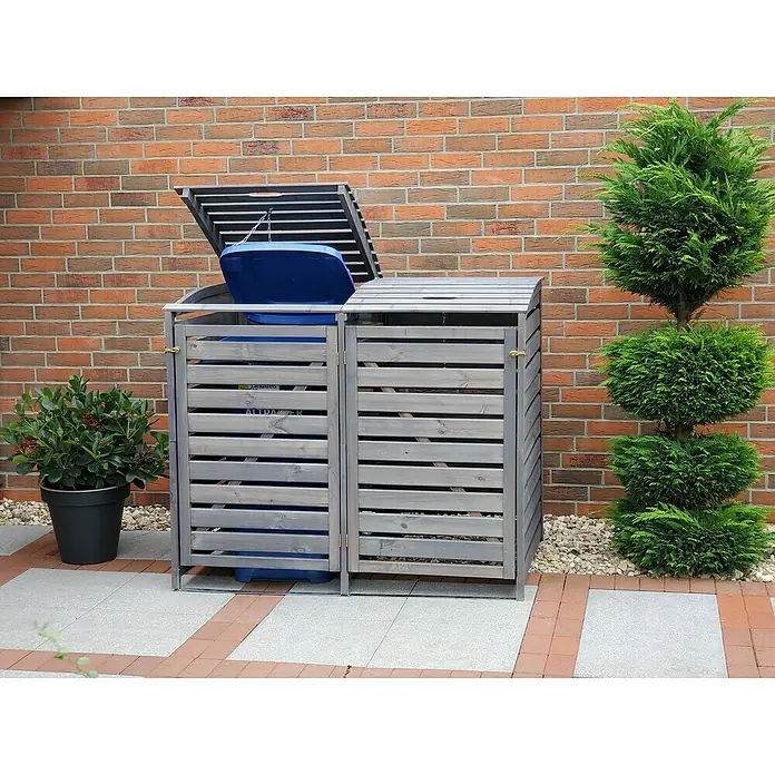 Promadino Mülltonnenbox Vario III -  - Diese Aufnahme zeigt eine graue Mülltonnenbox mit einer teilweise geöffneten Klappe, die zwei blaue Mülltonnen verbirgt, neben einem akkurat geschnittenen Zypressenbaum.