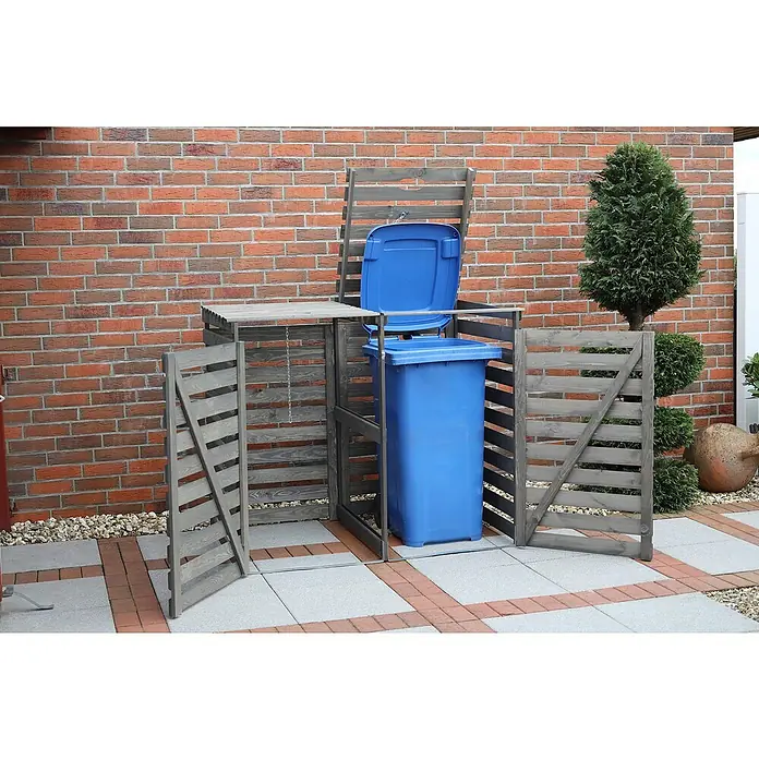 Promadino Mülltonnenbox Vario V -  - Das Bild zeigt eine Mülltonnenbox aus Holz mit einer blauen Mülltonne darin, vor einer Ziegelwand auf einer gepflasterten Fläche.