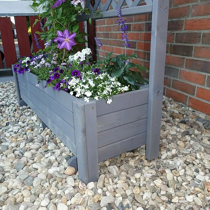 Promadino Pflanzkasten Romantica - Außenmaß (L x B x H): 210 x 38 x 80 cm, Holz, Grau - Nahaufnahme eines bepflanzten, grauen Blumenkastens mit Spalier vor einem Ziegelhaus, umgeben von Kies.