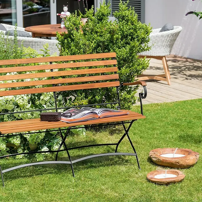 Sunfun Moni Klappbank - B x T x H: 120 x 57 x 90 cm, Akazie, Naturbraun/Schwarz, Klappbar - Eine hölzerne Gartenbank mit einem Buch und zwei Kerzen auf dem Rasen.