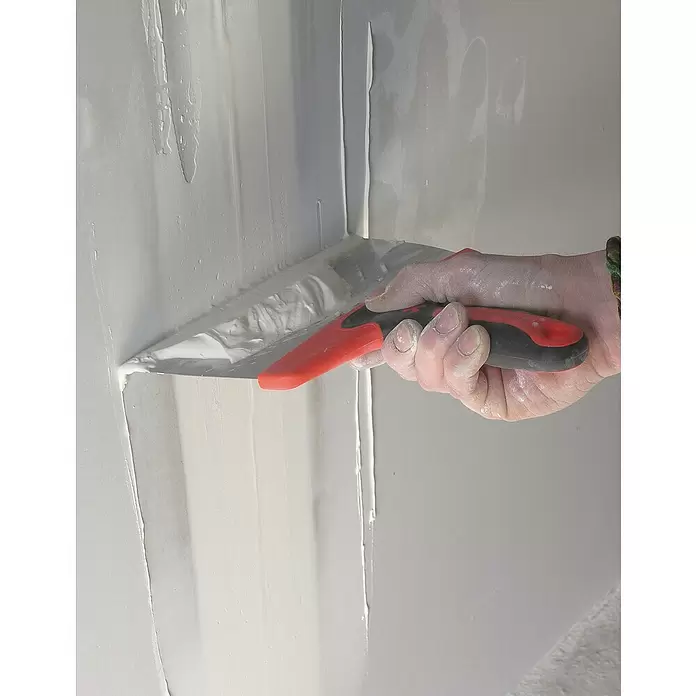 Mondelin Flächenspachtel - Breite: 240 mm - Hand hält eine rot-schwarze Spachtel und trägt weißen Spachtelmasse auf eine Wandecke auf.