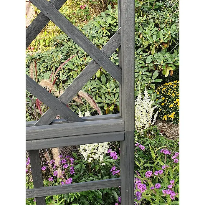 Promadino Pergola Ben - 60 x 200 x 210 cm, Anthrazit, Holz - Detailaufnahme eines grauen Holzzauns im Garten, umgeben von üppigen Pflanzen und bunten Blumen, die eine natürliche und friedliche Atmosphäre schaffen.