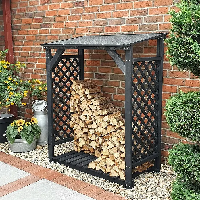Promadino Kaminholzregal Speyer - 120 x 68 x 148 cm, Holz, Anthrazit - Ein mit Holz gefüllter, dunkler Holzschuppen steht vor einer Backsteinmauer, umgeben von Pflanzen.
