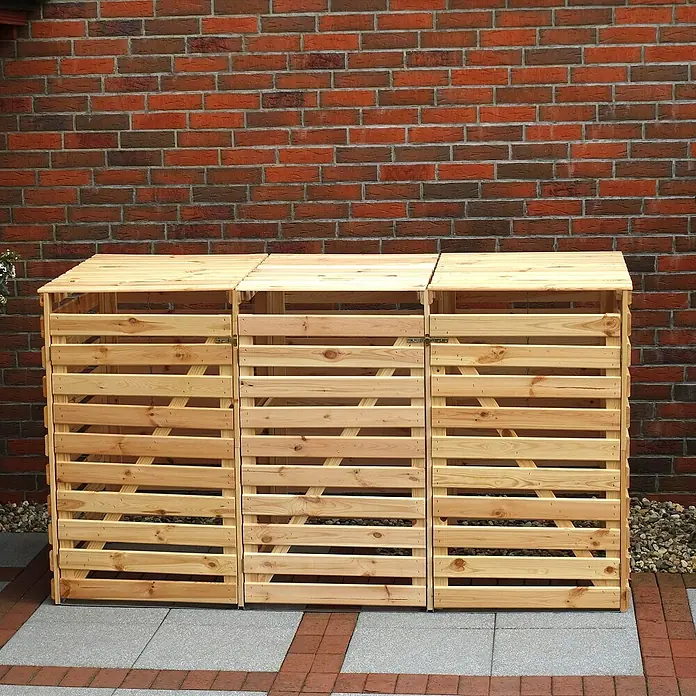 Promadino Mülltonnenbox Variov -  - Eine hölzerne Mülltonnenbox mit drei Fächern vor einer roten Backsteinmauer. Die Box ist aus hellem Holz und hat Lamellen.
