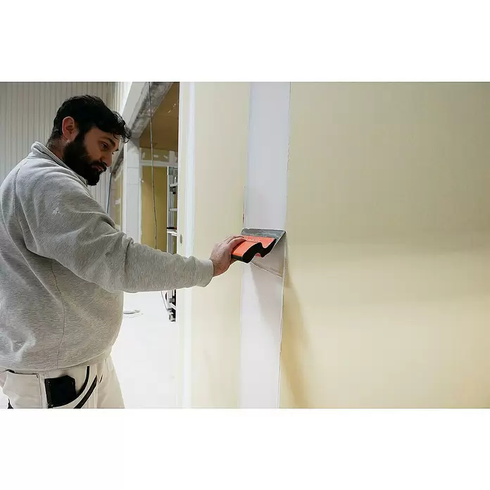 Mondelin Feinspachtel Ergolame - L x B: 250 x 100 mm, Stärke: 0,6 mm - Mann glättet Spachtelmasse mit einer orange-silbernen Spachtel an einer Wand.