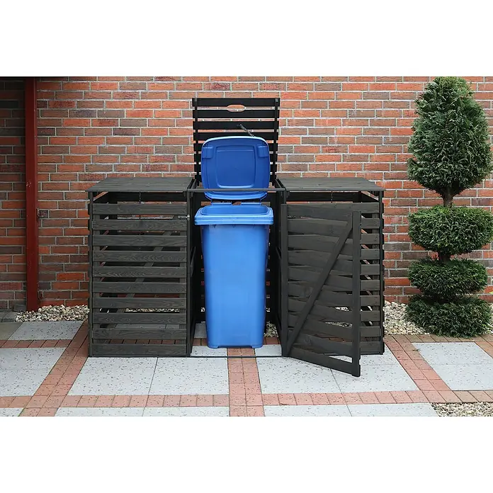 Promadino Mülltonnenbox Variov -  - Das Bild zeigt eine graue Mülltonnenbox mit einer blauen Mülltonne darin, die im Garten vor einer Backsteinmauer steht.
