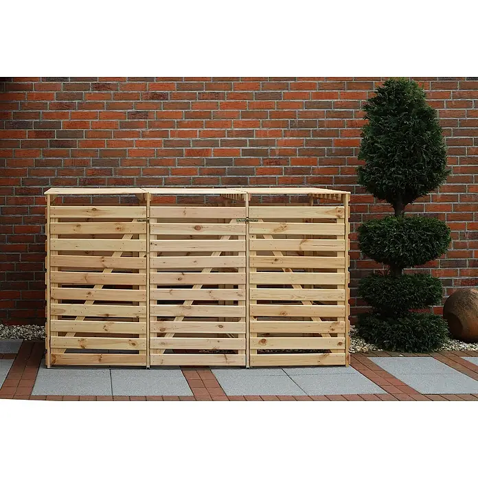 Promadino Mülltonnenbox Variov -  - Drei hölzerne Mülltonnenboxen stehen vor einer roten Backsteinmauer und einem grünen, gestutzten Baum. Sie bieten eine stilvolle Verkleidung für Mülltonnen.