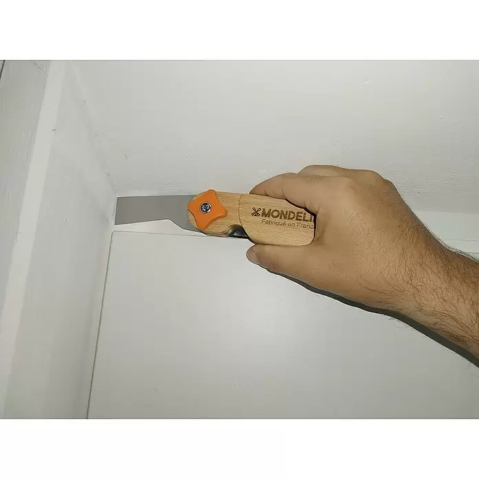 Mondelin Multifunktions-Messer Heliss - Ausstattung: 4 Klingenpositionen, Holz - Hand hält ein Mondelin Cuttermesser mit Holzgriff und orangefarbener Arretierung, Klinge ritzt eine weiße Wandecke.