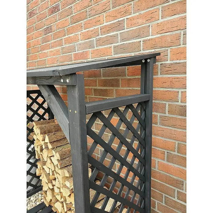 Promadino Kaminholzregal Speyer - 120 x 68 x 148 cm, Holz, Anthrazit - Das Bild zeigt ein schwarzes Holzlager mit Gittermuster, gefüllt mit Holzscheiten, vor einer Backsteinmauer.