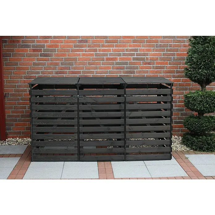 Promadino Mülltonnenbox Variov -  - Ein dreifacher, anthrazitfarbener Mülltonnenbox aus Holz steht vor einer Backsteinmauer. Rechts daneben steht ein grüner Busch.