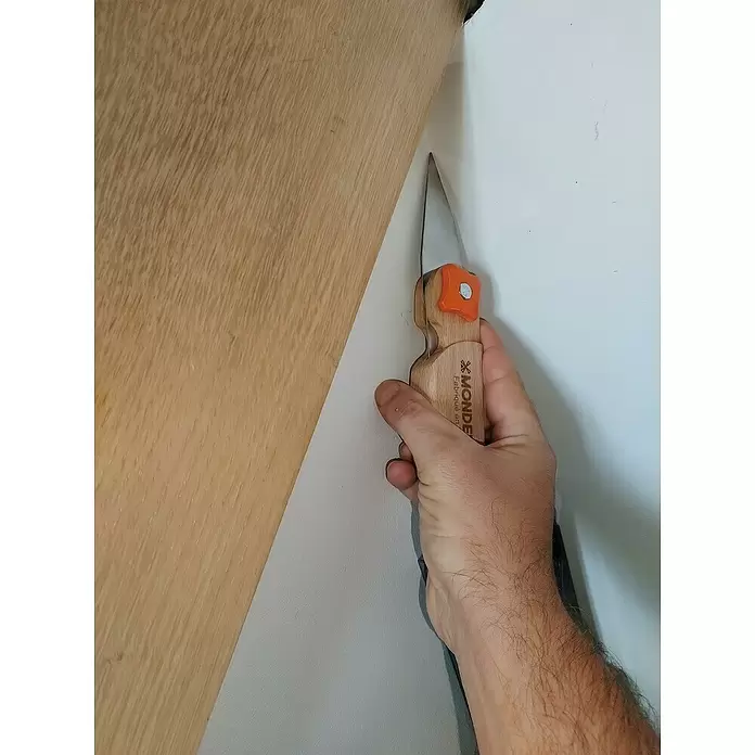 Mondelin Multifunktions-Messer Heliss - Ausstattung: 4 Klingenpositionen, Holz - Eine Hand verwendet ein Schneidemesser mit Holzgriff und orangefarbenem Akzent, um weißes Material an einer Ecke neben einer hellen Holzoberfläche zu schneiden.