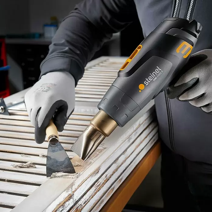 Steinel Heißluftgebläse HM 1920 E - 2.300 W, Temperaturbereich: 80 °C - 630 °C - Behandschuhte Person entfernt mit schwarzem Steinel HM 1920 E Heißluftgebläse und Schaber alte Farbe von einem weißen Holzfensterladen.