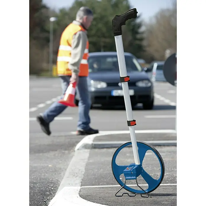 Bosch Professional Messrad GWM 32 - Messbereich: Max. 9999,99 m, Messgenauigkeit: 10 cm/100 m - Ein blaues Rollmessrad steht auf einer Straße, ein Mann im Hintergrund trägt eine Warnweste und einen Verkehrskegel.
