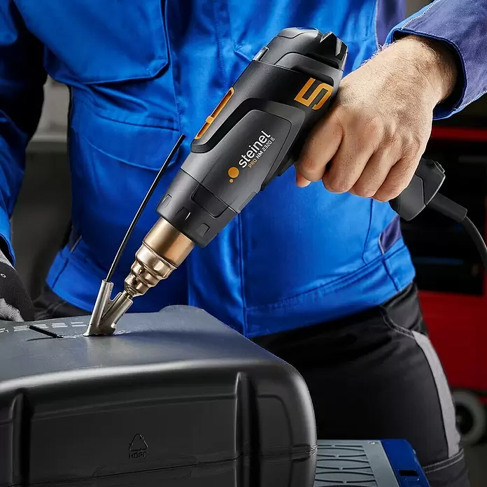 Steinel Heißluftgebläse HM 2320 E KF - 2.300 W, Temperaturbereich: 80 °C - 650 °C - Person nutzt eine schwarz-gelbe Steinel Heißluftpistole mit Schweißaufsatz an einem schwarzen Kunststoffteil.