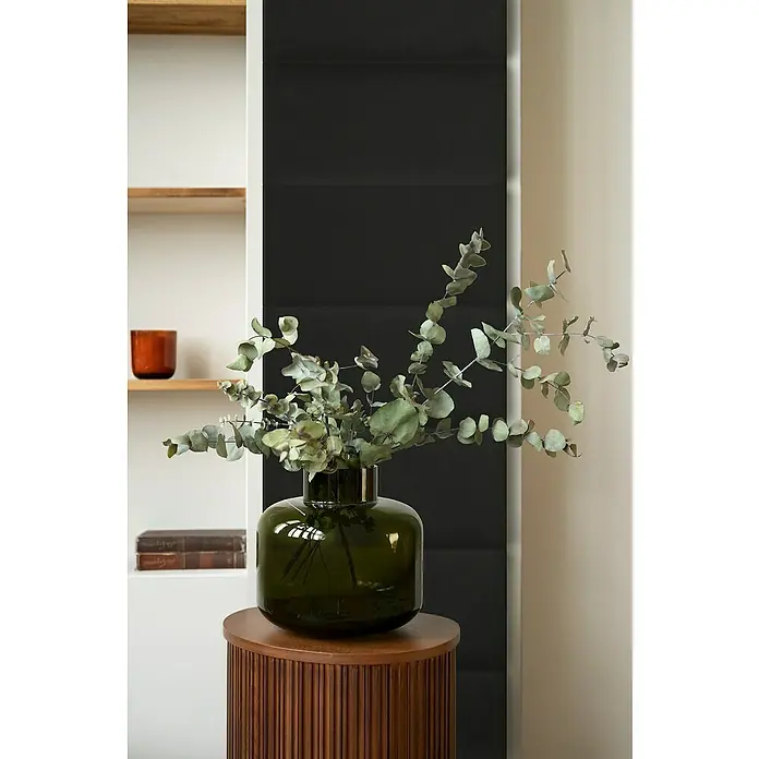 Fllow Wandpolster Velvet - Schwarz, L x B: 60 x 30 cm - Ein grünes Glasgefäß mit Eukalyptuszweigen steht auf einem braunen Beistelltisch, vor einem Regal. Das minimalistische Design sorgt für Eleganz.
