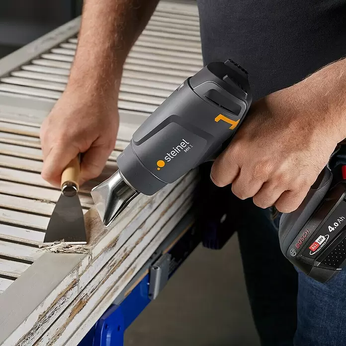 Steinel Akku-Heißluftgebläse MobileHeat 1 - 1 Akku, Temperaturbereich: 300 °C - 450 °C - Eine Person entfernt mit einem Steinel MH 1 Akku-Heißluftgebläse und einem Spachtel Farbe von einer Holzjalousie. Betrieben mit einem Bosch 18V 4.0Ah Akku.