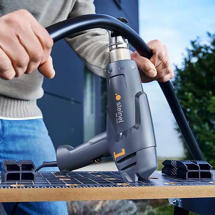 Steinel Heißluftgebläse HM 1620 S - 2.000 W, Temperaturbereich: 80 °C - 600 °C - Graues Steinel Heißluftgebläse mit orangefarbenen Akzenten erhitzt ein schwarzes Rohr, von einer Person an einem Arbeitstisch gehalten.
