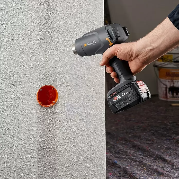 Steinel Akku-Heißluftgebläse MobileHeat 1 - 1 Akku, Temperaturbereich: 300 °C - 450 °C - Hand hält eine graue Steineil Heißluftpistole mit Bosch 18V Akku, gerichtet auf eine strukturierte weiße Wand neben einer orangen Elektrobox.
