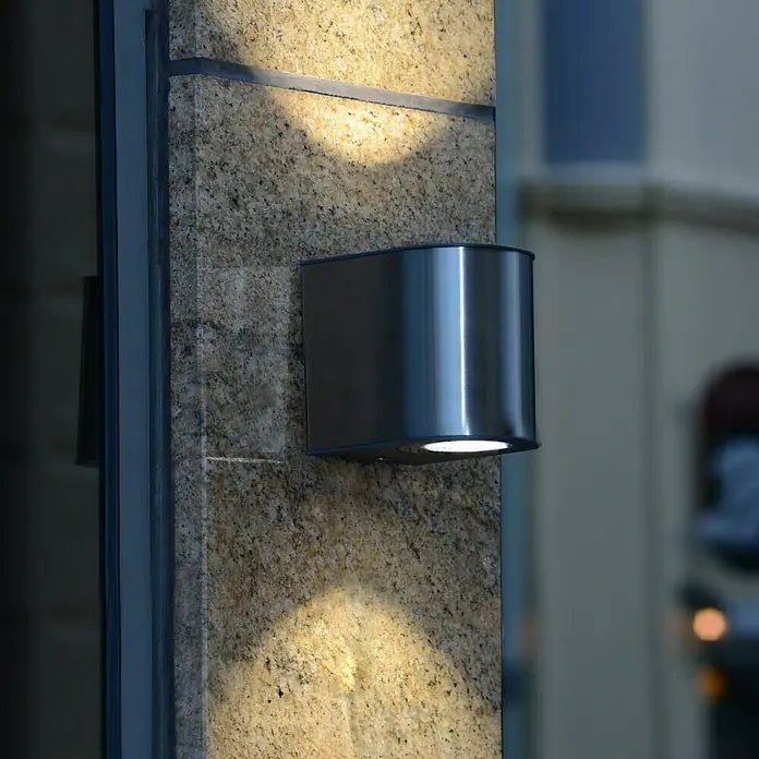 Lutec Aplique exterior LED Gemini - 24 W, 13,5 x 14,1 x 10,6 cm, Acero inoxidable, IP54 - Primer plano de una lámpara de pared cilíndrica moderna de metal, que proyecta una luz cálida sobre una superficie de piedra estampada.