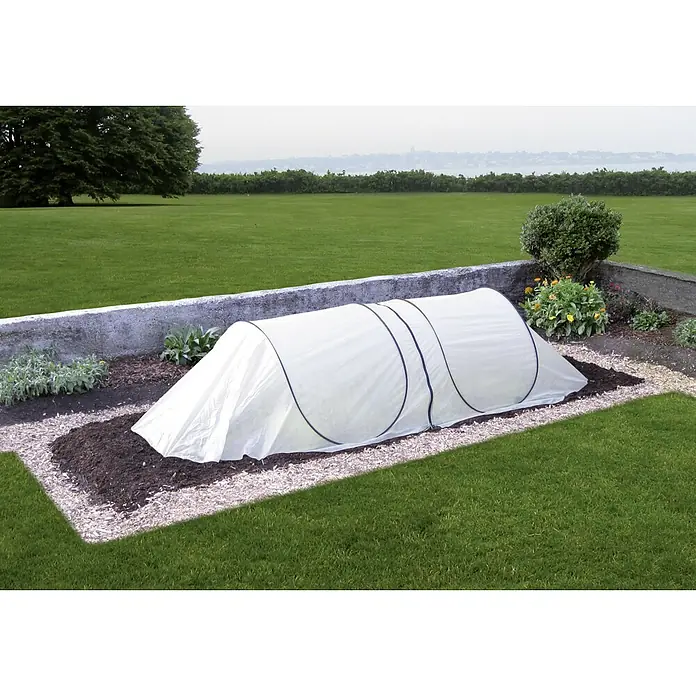 GardenGuard Kälteschutz - 250 x 80 x 60 cm, Weiß, Vlies - Ein Garten mit einem weißen Gewächshaus-Tunnel, der Pflanzen vor Witterungseinflüssen schützt, eingebettet in eine gepflegte Rasenfläche.