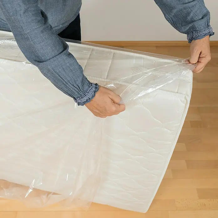 Matratzenhülle - 240 x 130 cm, Transparent, 2 Stk. - Eine Person packt eine neue Matratze aus einer Plastikfolie aus, bereit für den Gebrauch in einem Schlafzimmer.