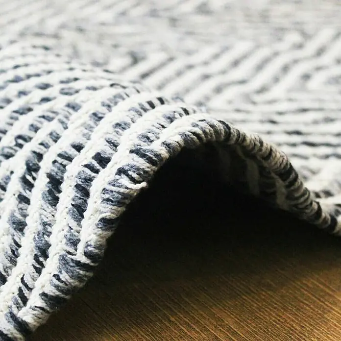 Kayoom Flachgewebeteppich Cocktail 400 - Grau, 170 x 120 cm, 100% Baumwolle - Nahaufnahme eines grauen und weißen Teppichs mit einem gewebten Zickzackmuster. Der Teppich liegt auf einem Holzboden.
