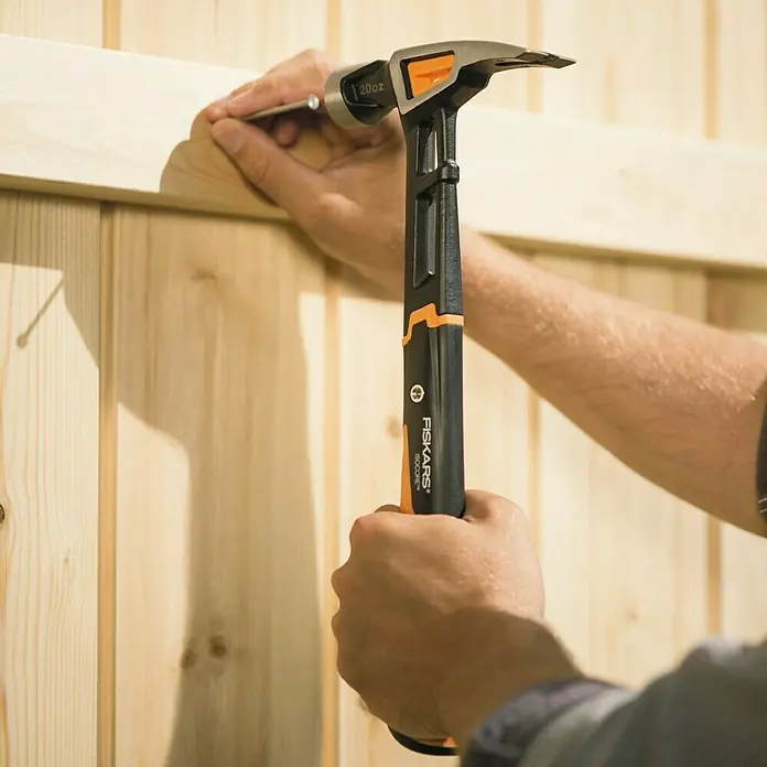 Fiskars Universalhammer XL - Gewicht Kopf: 570 g, Länge: 390 mm - Nahaufnahme eines Mannes, der mit einem Fiskars-Hammer einen Nagel in ein Holzstück schlägt. Der Hammer ist schwarz mit orangefarbenen Akzenten.