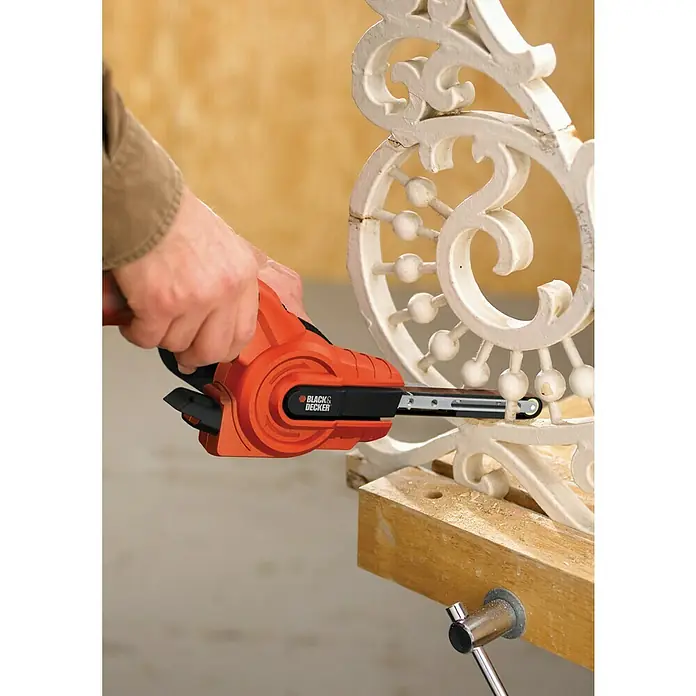 Black+Decker Lijadora de banda estrecha eléctrica KA900E - 350 W - Primer plano de una herramienta eléctrica BLACK+DECKER que se utiliza para lijar una pieza de madera blanca ornamentada.