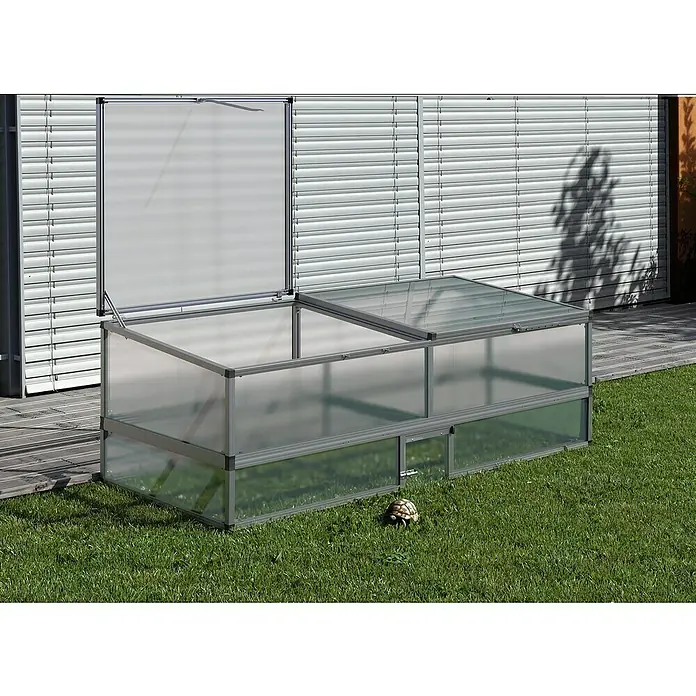 KGT Freilaufgehege - 205 x 91 x 26 cm, Aluminium, Pressblank - Nahaufnahme eines Gewächshauses mit geöffnetem Deckel auf einer Wiese, neben einer Hauswand mit Jalousien.