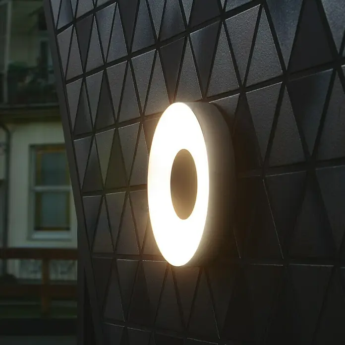 Lutec Aplique exterior LED Ublo - 7,5 W, 4,5 x 19,3 x 19,3 cm, Plateado, Opal, IP54 - Una lámpara circular que emite un brillo cálido, montada en una pared con patrones triangulares. El diseño moderno enfatiza la estética geométrica.