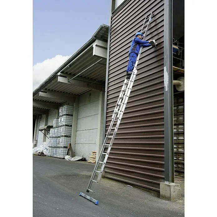 Krause Stabilo Sprossen-SeilzugleiterArbeitshöhe: 8,45 m, 30 Stk., Aluminium - Ein Handwerker auf einer ausziehbaren Leiter, der an der Fassade eines Lagerhauses arbeitet. Fokus auf die vielseitige Leiter für Profis.