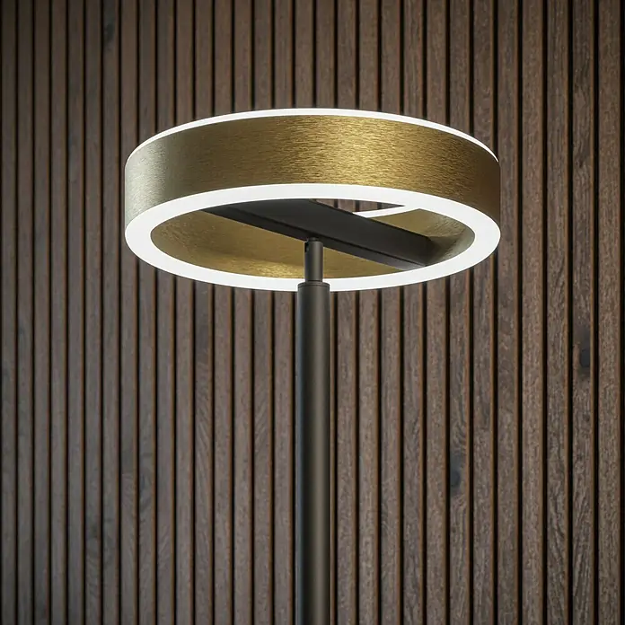 Tween Light LED-Stehleuchte Orlando - Höhe: 180 cm, Sandschwarz, Messing gebürstet, Warmweiß - Nahaufnahme einer Stehlampe mit einem goldenen, ringförmigen Schirm und einem schlanken, schwarzen Ständer vor einer Holzwand.