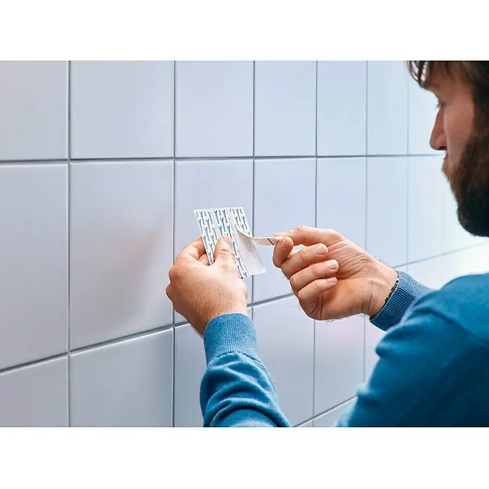Tesa Clavo adhesivo ajustable - Blanco, Carga soportada: 4 kg, 2 ud. - Una persona retira la película protectora de una tira adhesiva Tesa Powerstrips para adherirla a azulejos.