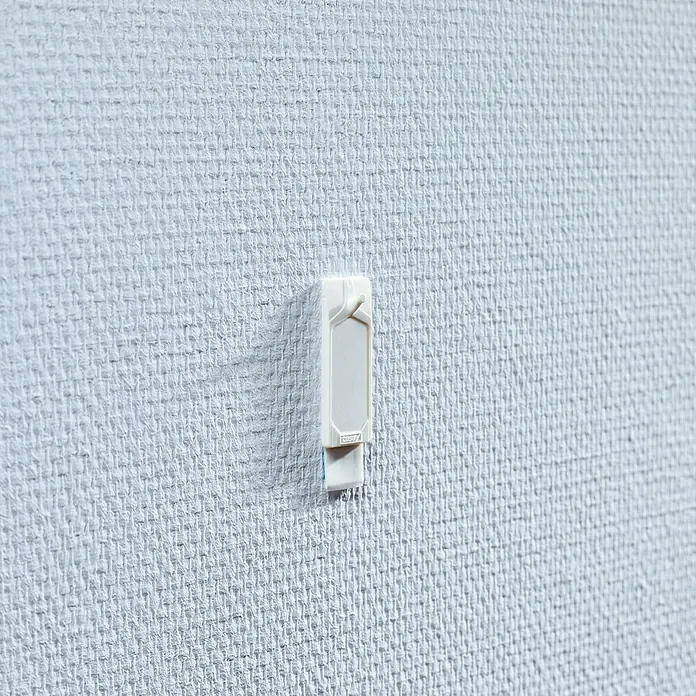 Tesa Clavo adhesivo - Blanco, Carga soportada: 0,5 kg, 2 ud., Paredes pintadas - Primer plano de un clip de cable autoadhesivo blanco fijado a una pared gris claro con textura. Gestión práctica de cables en casa.