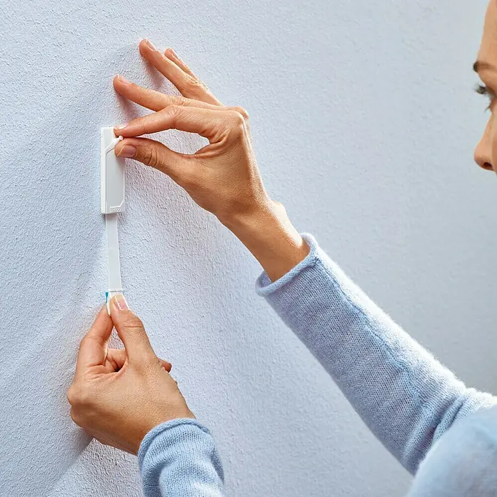 Tesa Clavo adhesivo - Apto para: Paredes pintadas, Carga soportada: 1 kg, 2 ud., Blanco - Una mujer sujeta una tira adhesiva Tesa blanca a una pared clara, para colgar algo.