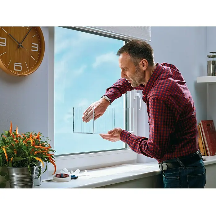 Tesa Powerbond Cinta de doble cara Vidrio - 1,9 cm x 5 m, Transparente - Un hombre usa una película adhesiva transparente para fijar aislamiento adicional a una ventana, buscando mejorar la eficiencia energética.