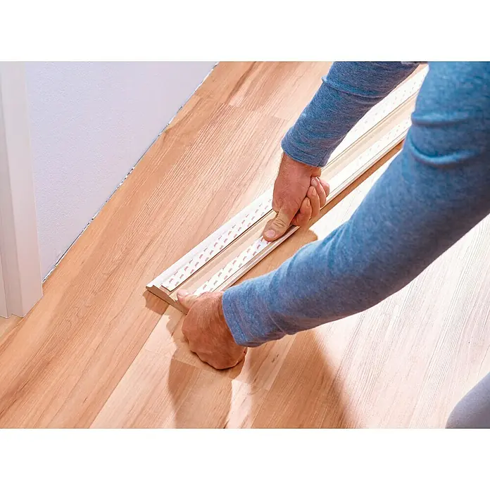 Tesa Powerbond Cinta de doble cara Paredes pintadas/Yeso - 1,9 cm x 1,5 m, Blanco - Una persona utiliza tiras adhesivas para fijar un zócalo a un suelo de madera, creando una transición perfecta.