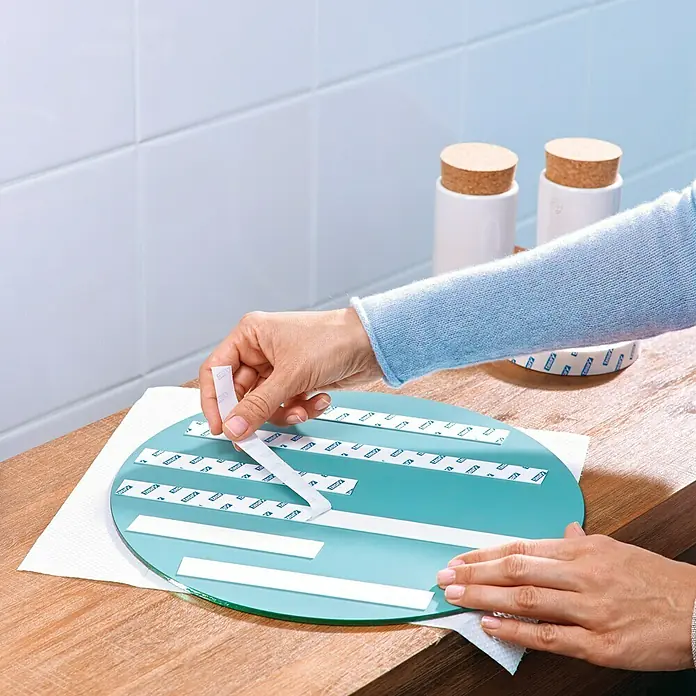 Tesa Powerbond Cinta de doble cara Resistente humedad - 1,9 cm x 1,5 m, Blanco - Una persona aplica cinta de montaje de espejo a un espejo redondo. Al fondo, dos recipientes con tapones de corcho sobre una tabla de madera.