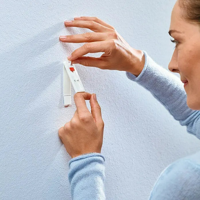 Tesa Clavo adhesivo ajustable - Blanco, Carga soportada: 1 kg, 2 ud. - Primer plano de una mujer que fija un clavo adhesivo a una pared blanca. El clavo adhesivo innovador ofrece una solución práctica.