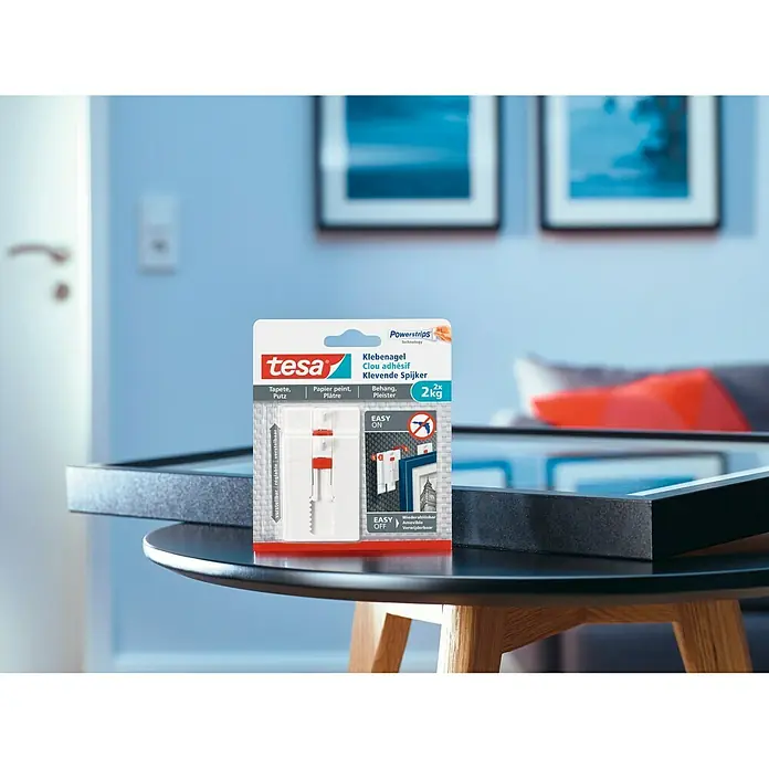 Tesa Clavo adhesivo ajustable - Blanco, Carga soportada: 2 kg, 2 ud. - Primer plano del producto Tesa Klebenagel Clou adhésif Klevende Spijker sobre una mesa, con una pared azul al fondo.