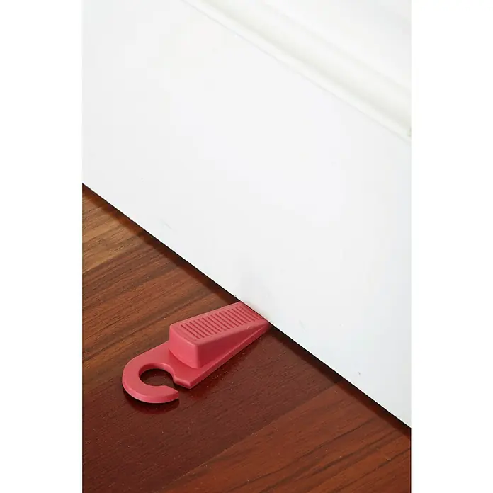 Arregui Tope para puertas A-1044290 - Plástico, 1 ud. - Primer plano de una cuña de puerta de plástico roja que mantiene una puerta blanca abierta sobre un suelo de madera oscura, con un gancho para colgar el tope de puerta.