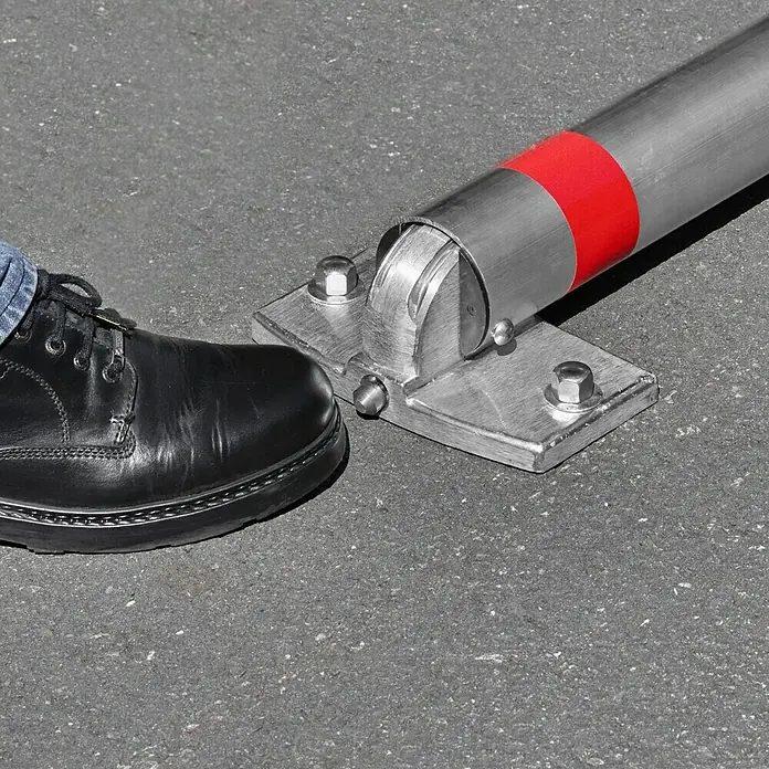 Urbanus Sperrpfosten - Höhe über Boden: 900 mm, Durchmesser: 76 mm, Aluminium, Profilzylinderschloss, Montageart: Aufdübeln - Detailaufnahme einer umklappbaren Parkplatzsperre aus Edelstahl mit rotem Band und schwarzem Lederschuh auf Asphalt.