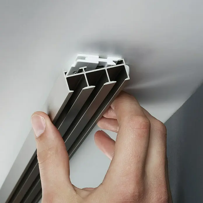 Expo Ambiente Gardinenprofil Smart Set - Anzahl Läufe: 3 Stk., Silber, 160 cm - Eine Hand hält eine braune Gardinenschiene mit weißer Befestigung, die an einer weißen Decke montiert wird. Detailaufnahme.