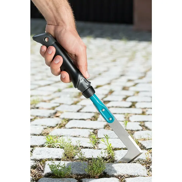 Gardena Comfort Hand-Fugenkratzer - Stahl - Nahaufnahme eines Mannes, der einen Fugenkratzer mit blauem Griff verwendet, um Unkraut zwischen Pflastersteinen zu entfernen.