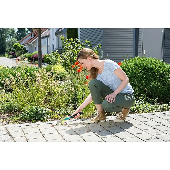 Gardena Comfort Hand-Fugenkratzer - Stahl - Eine Frau jätet Unkraut mit einem Fugenkratzer auf einem gepflasterten Weg. Das Werkzeug verfügt über einen ergonomischen Griff.