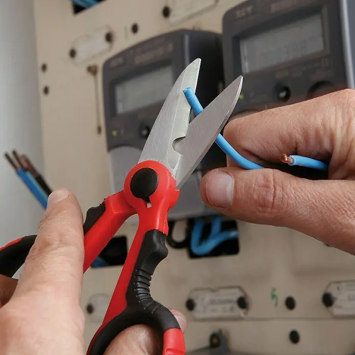 Super Ego Tijeras de trabajo para electricistas - Largo: 14,4 cm, Longitud de corte: 42 mm, Rojo/Negro - Primer plano de unas tijeras con mangos rojos cortando un cable eléctrico azul, con un panel eléctrico de fondo.