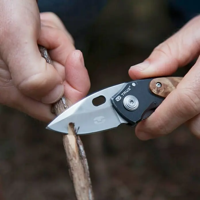True Utility Taschenmesser Jacknife TU576K - Edelstahl 420 - Nahaufnahme eines TRUE-Taschenmessers, das einen kleinen Stock bearbeitet, wodurch die Vielseitigkeit des Werkzeugs hervorgehoben wird.