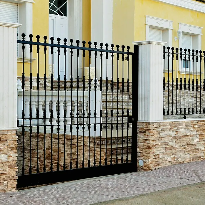 Revestimiento de piedra Petra Arena - Cantidad total: 4 pzs., Arena - Esta imagen muestra una elegante puerta de hierro forjado negro con detalles decorativos, ubicada en una pared de piedra con pilares y una casa amarilla.