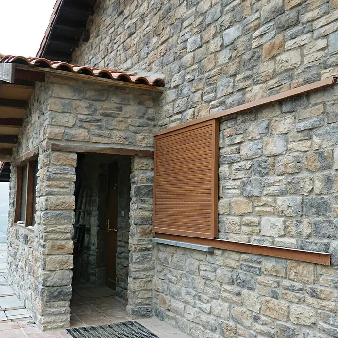 Revestimiento de piedra Silleria Plomo - Gris, Cantidad total: 4 pzs. - Primer plano de una casa con pared de piedra y contraventana de madera parcialmente abierta. La casa tiene un tejado de tejas y tiene un aspecto rústico.